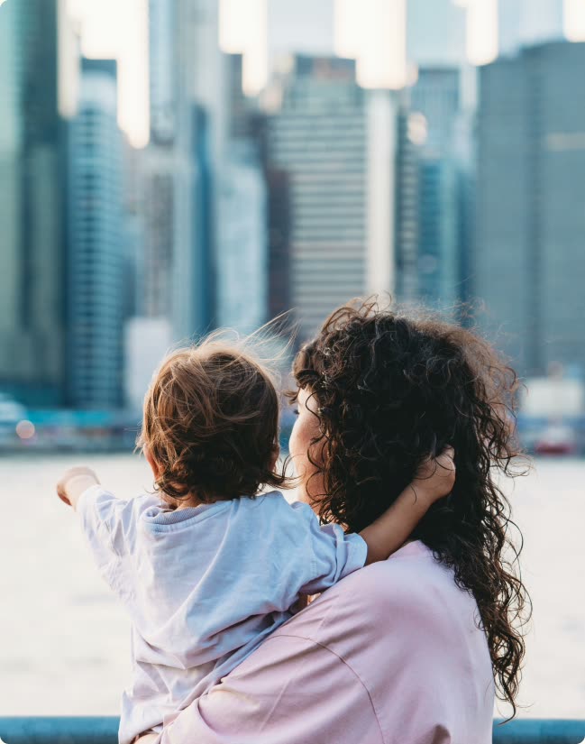 A woman holding a young child, both looking out across the water toward the Manhattan skyline.